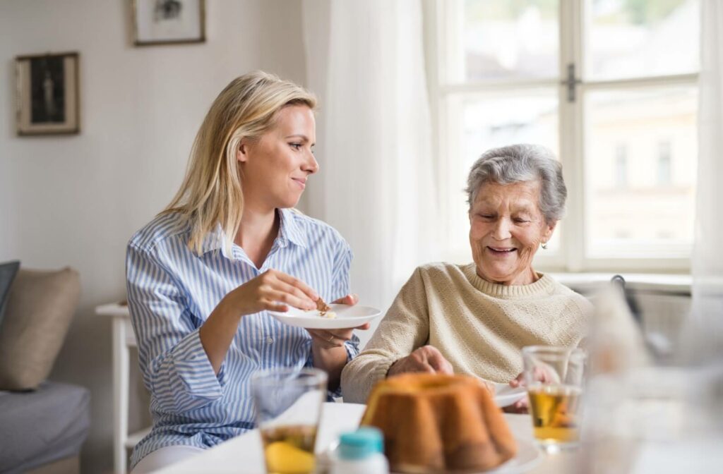 A senior woman discusses the activities she's involved in at her assisted living community with her daughter while they share a snack.