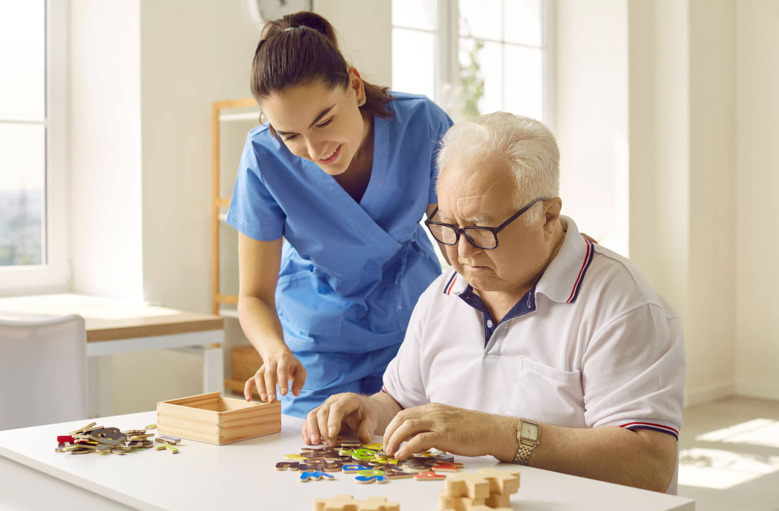 A smiling caregiver helping an older adult with dementia sort through puzzle pieces in memory care.