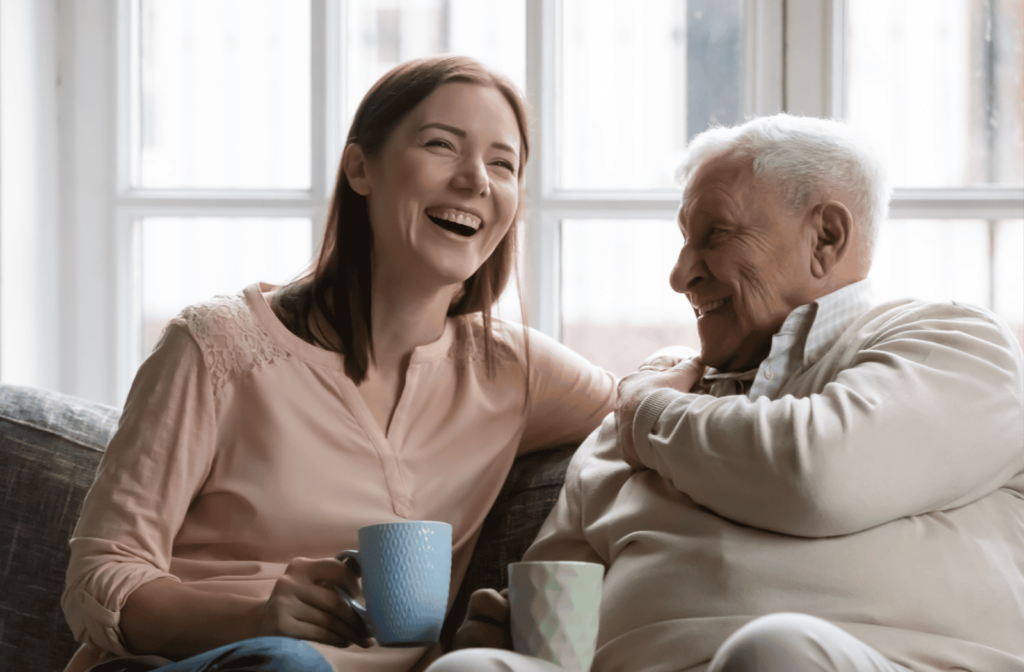 Senior parent and adult child sit on a couch with coffee laughing.