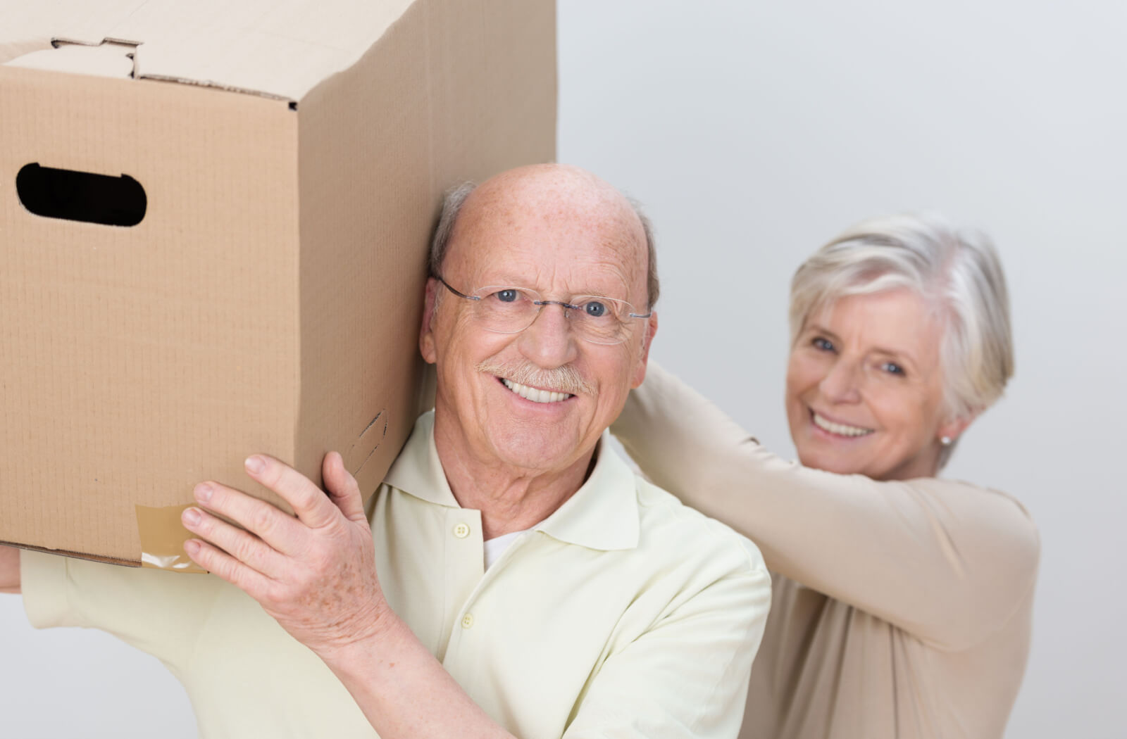 An older couple smiling and working together to carry a box when downsizing for a move to assisted living.