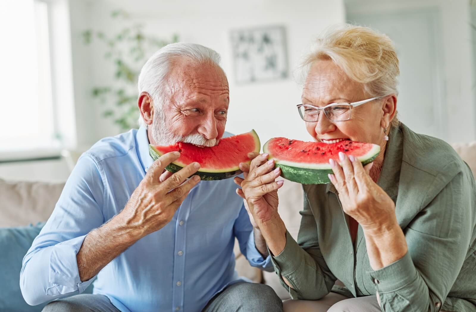 Two older adults smiling and eating large watermelon slices together indoors enjoying a refreshing summer snack