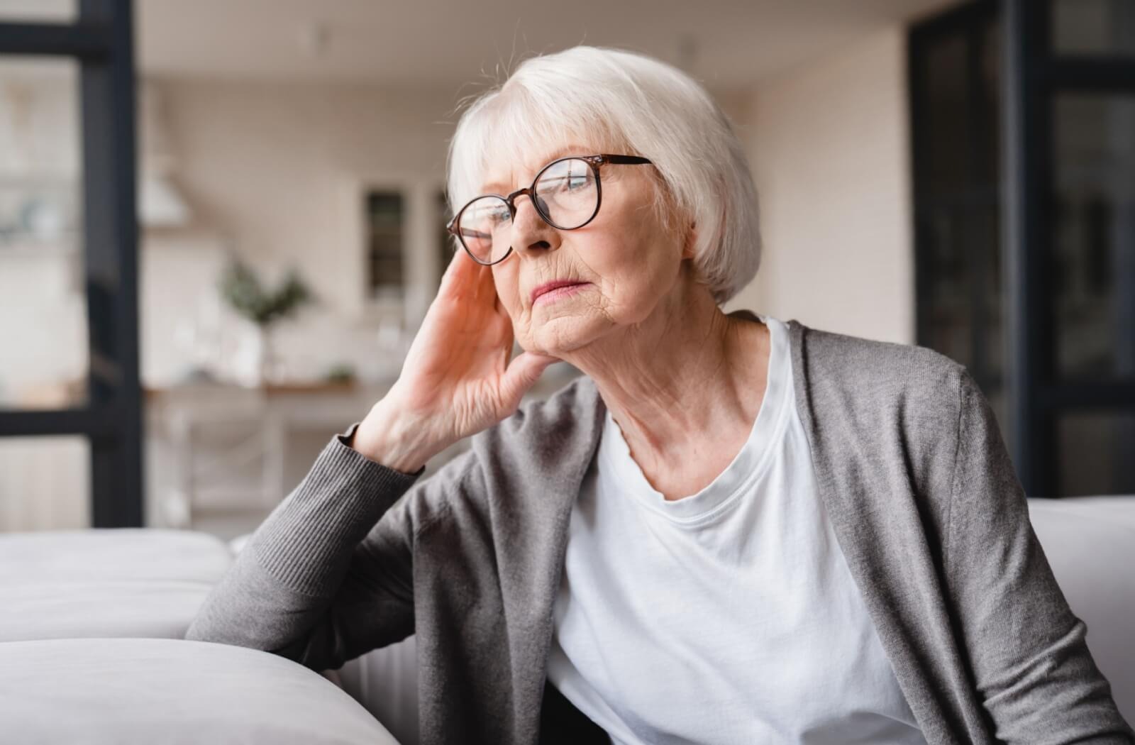 A senior sits on a couch and looks into the foreground while thinking about senior living options.