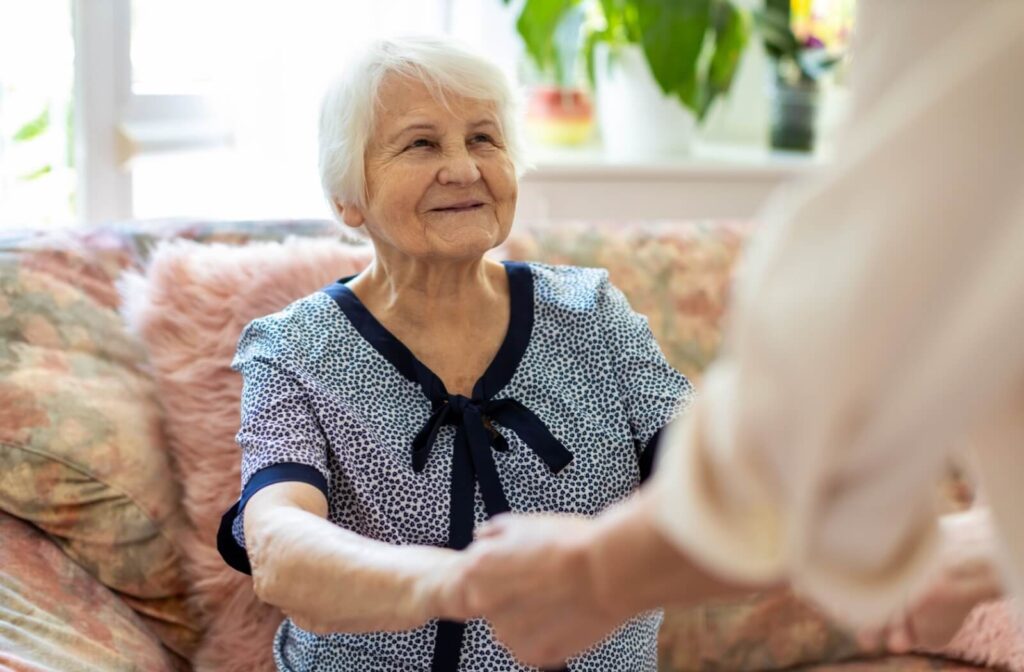 A caregiver holds hands with a seated, smiling older adult in a bright memory care setting.